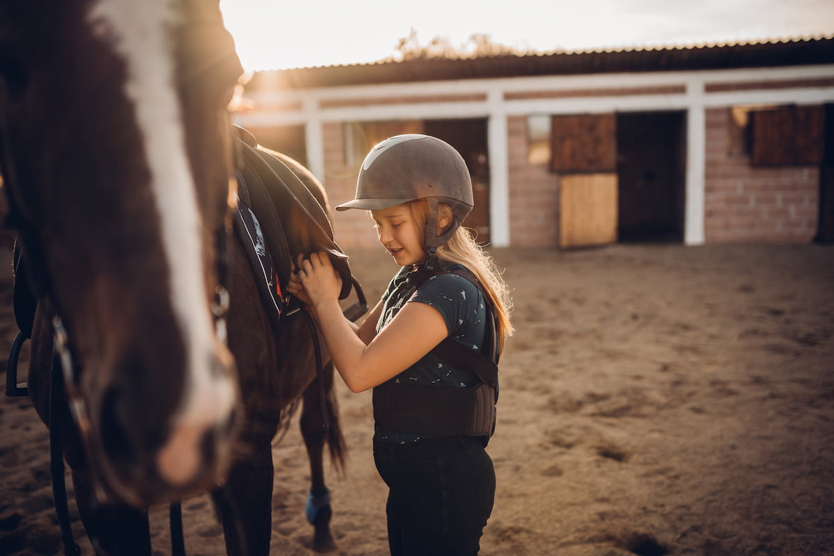 Kind mit Sicherheitsweste und Reithelm beim Satteln eines Pferdes vor einem Reitstall