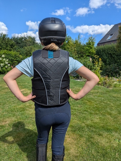 Kind mit Reithelm und Schutzweste von hinten, bereit für den Reitunterricht